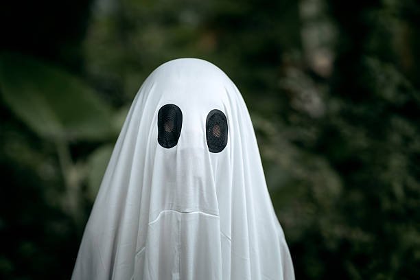 A young boy dressed as a ghost for Halloween wears a white sheet with two black eye holes. He raises both arms in a spooky gesture, surrounded by dark green foliage