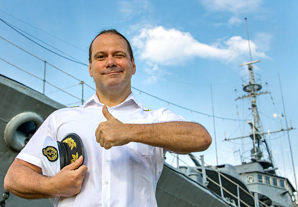 Captain standing in dock before warship and  shows thumb up. A sailor officer in white uniform stands beside battleship.