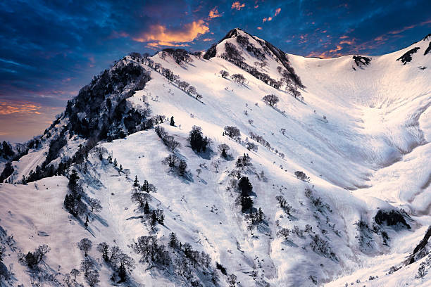A stunning winter mountain landscape illuminated by warm sunset light. Snow-covered slopes, scattered trees, and dramatic colorful clouds create a breathtaking alpine scene. Perfect for themes of travel, adventure, nature, wilderness, winter holidays, scenic beauty, exploration, and outdoor freedom.