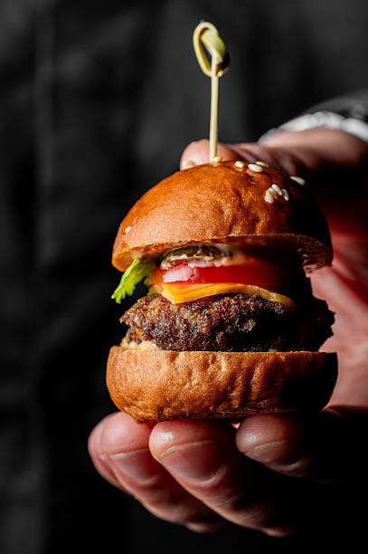 istock A small cheeseburger slider, held in a person's hand, features a grilled patty, melted cheese, fresh tomato, and lettuce on a sesame seed bun, against a dark background 2243983965