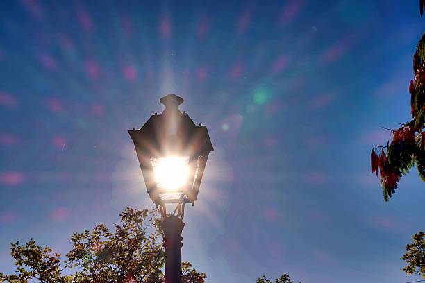 A vintage lamp post stands tall against a vibrant blue sky, with sunlight illuminating it. Rays of light create a striking lens flare, showcasing a bright midday scene.