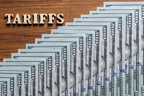 Wooden letters with the inscription tariffs and $100 American paper bills on table. Tariffs overlaid on US dollars, symbolizing trade wars and tariff policies during Trump administration
