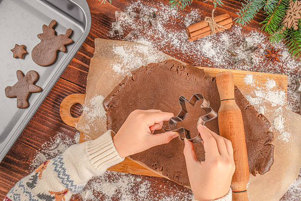 istock Hands cutting gingerbread dough with cookie cutter on wooden board, Christmas baking process with flour, rolling pin, cinnamon sticks, fir branches, and baking tray, cozy holiday atmosphere, top view. 2244008198