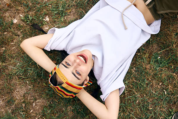 A cheerful young woman relaxing on the grass, wearing a colorful cap and casual attire, enjoying the outdoors. Her vibrant mood and relaxed posture capture a youthful and playful spirit.