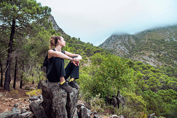 A young Caucasian woman sits on a large rock after a hike, taking a break and enjoying the panoramic view of the vast green landscape and cloudy sky in the mountains near Ojen, Malaga. Concept of well-being and personal achievement.