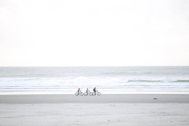 A woman goes for a bike ride at low tide on Long Beach on Vancouver Island near Tofino, British Columbia, Canada, at sunset. She rides a mountain bike and wears casual clothing and a sun hat.
