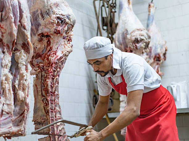 istock Worker at a butcher's shop using a hook to hold the meat while cutting a block with a saw 2244032621