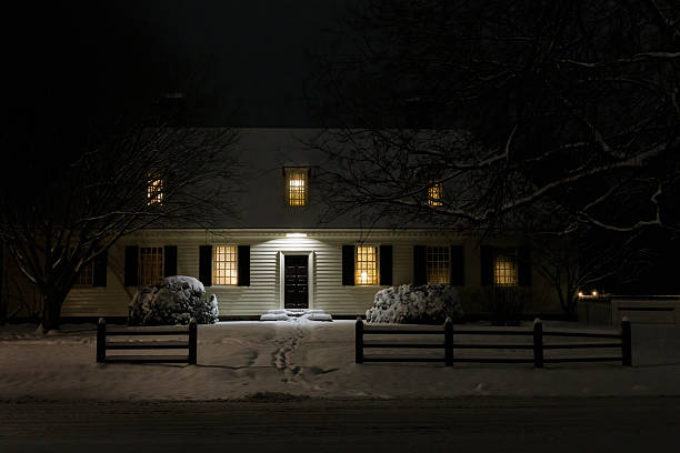 A picturesque wooden cabin covered in snow during a nighttime snowfall. The warm glow from inside and festive string lights on the balcony contrast beautifully with the cold, snowy surroundings. Snowflakes fall gently in the foreground, creating a magical and cozy winter atmosphere. A reindeer-shaped light decoration adds a festive touch to the scene.