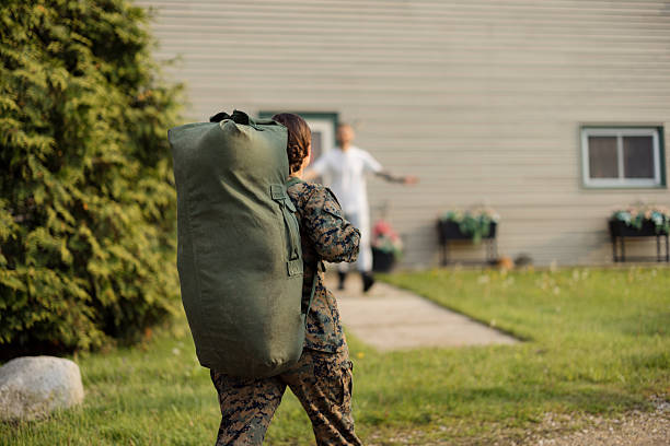 Military service member in camouflage carrying a duffel bag, walking towards an embracing family member outside a home