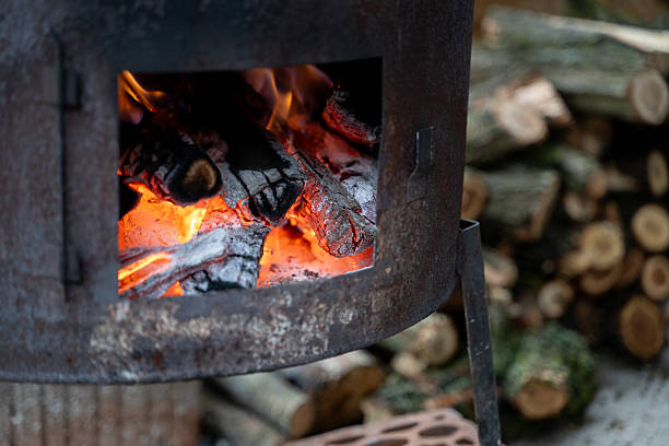 Close-up view of glowing embers and burning logs inside a rustic metal stove, with stacked firewood in the background. The vibrant orange flames and charred wood create a warm, intense atmosphere ideal for outdoor cooking or heating.