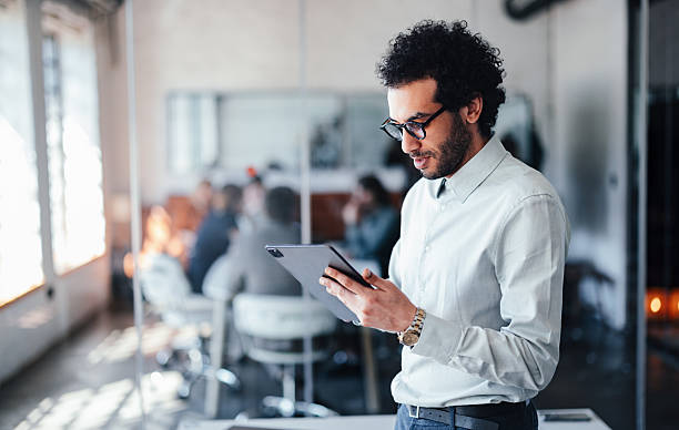 istock Professional Man Using Tablet in Modern Office While Colleagues Meet in Background During A Busy Team Meeting 2244066235