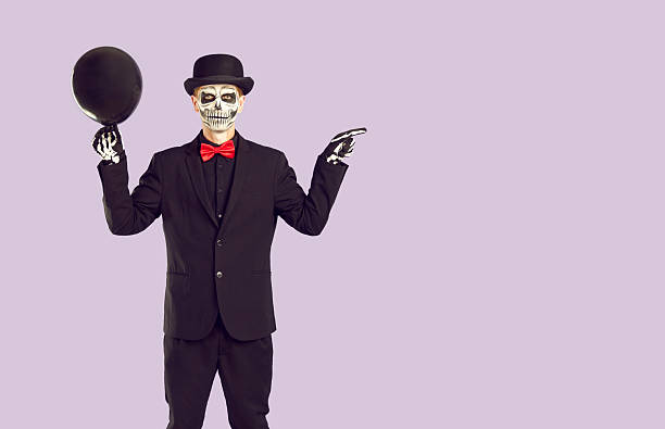 Skeleton man in black suit and hat, with skull makeup on face standing isolated on light purple background, holding black balloon and pointing finger at copy space on side. Halloween concept