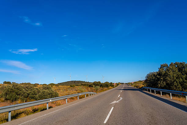 A winding road leads towards a distant hill dotted with wind turbines under a clear blue sky.