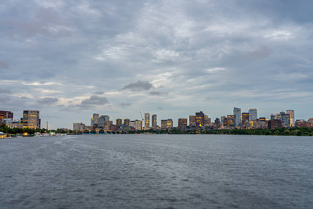 The Boston skyline is illuminated at dusk, with the Charles River flowing in the foreground and boats docked along the shore.