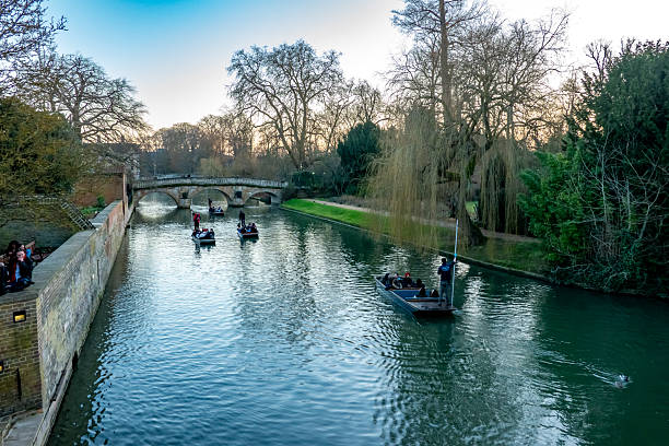 Cambridge, UK - February 02, 2025: Punting boats glide along the River Cam in Cambridge under an old stone bridge, surrounded by trees during winter season.