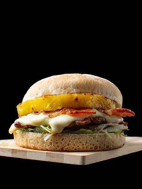 istock Gourmet Hawaiian Burger with Grilled Pineapple Slice, Bacon, and Melted White Cheese on a Wooden Board, Isolated on Black Background 2244077449