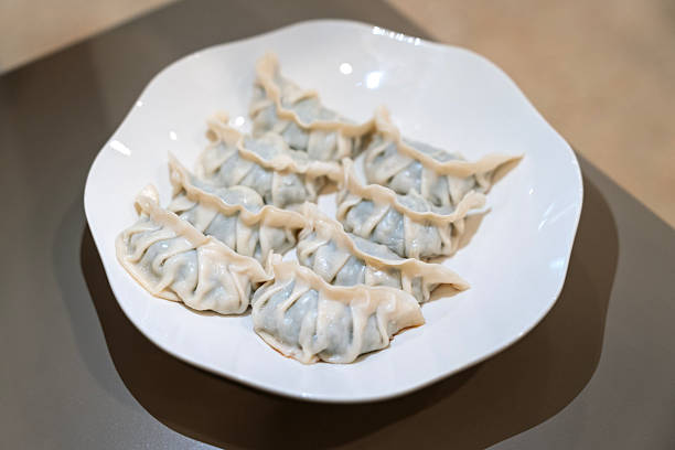 A close-up view of freshly steamed dumplings neatly arranged on a white plate. The dumplings feature soft, pleated wrappers filled with a savory mixture, showcasing Asian cuisine in a clean and minimal indoor dining setting.