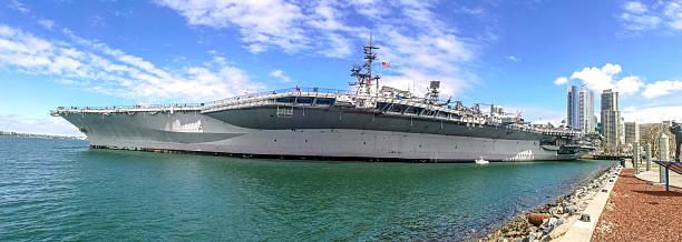 A large aircraft carrier is docked in the harbor with a clear blue sky overhead.