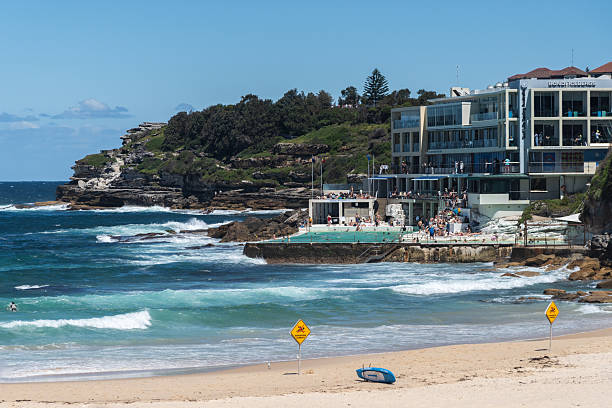 Bondi Beach, Sydney, Australia - February 16, 2025: Ocean waves roll in past Bondi Icebergs Swim Club, creating a white foam line. No people are on the sands along with two No Swimming sign.