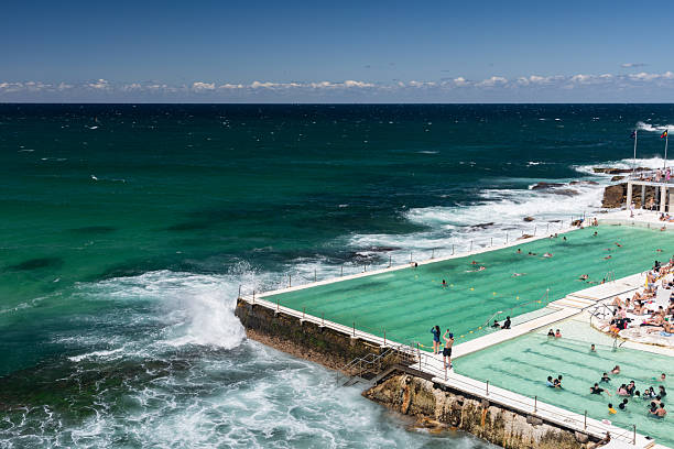 Bondi Beach, Sydney, Australia - February 16, 2025: The famous outdoor pool at the Icebergs Swim Club on Bondi Beach is seen here. People swim and sunbathe. Waves crash into the pool from the visible ocean floor, splashing water before disappearing.