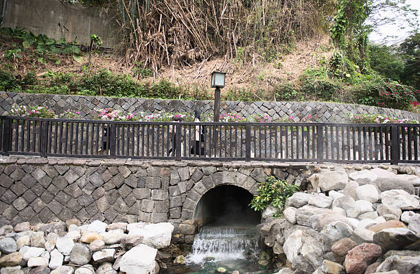 Xinbeitou Hot Spring area, Beitou, Taipei, Taiwan, Feb 27, 2024 : Hot spring water flows from a stone arch culvert into the creek below a wooden viewing platform, with a tourist walking by.