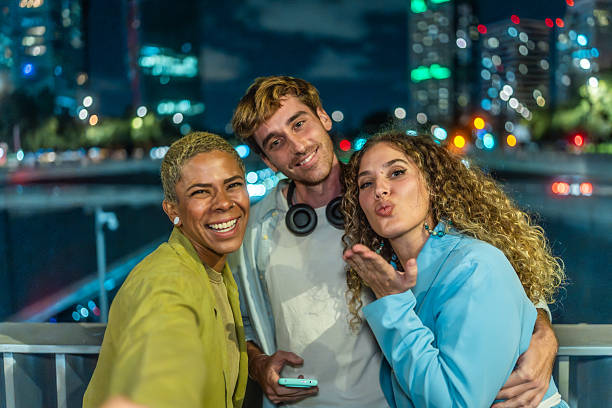 Three diverse adults are having fun at night in the city, smiling and blowing a kiss while posing together for a selfie with urban lights in the background