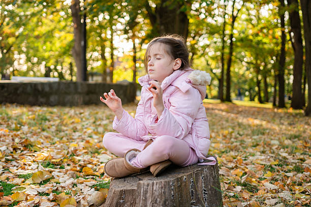 Young girl sitting in lotus position on a tree stump, practicing mindfulness and relaxation in nature during autumn