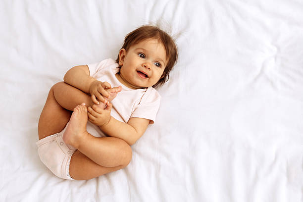 istock Cute baby lying on white bed and smiling while holding feet 2244113071