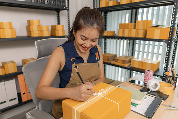 istock Young female entrepreneur writing a shipping address on a label for a parcel, preparing a package for delivery at her home office. She manages an online business and organizes shipments to customers. 2244117321