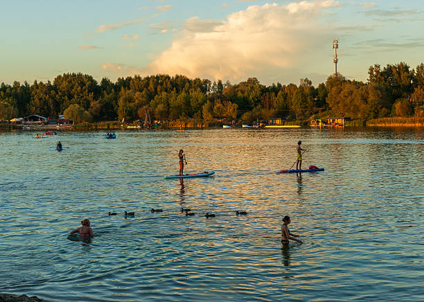 Krakow, Poland – September 21, 2025: Swimmers and paddleboarders enjoy the golden evening light on the calm lake, surrounded by forest and distant recreation areas. The water reflects the warm colors of sunset.