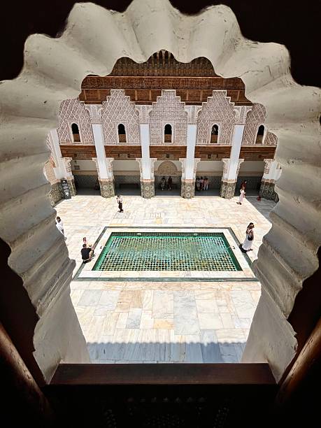 Ornate carved walls and wooden ceilings surround a central reflecting pool in the peaceful courtyard of Ben Youssef Madrasa, attracting tourists exploring historical Moroccan culture