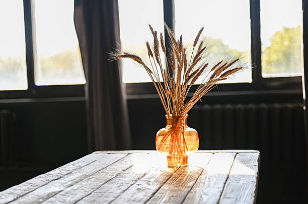 Dried wheat stalks standing in a translucent amber vase on a wooden table near a sunlit window