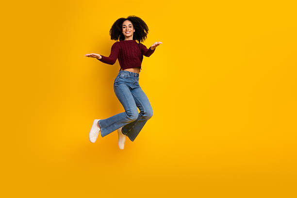 istock Young woman jumping in a bright yellow studio backdrop wearing a red sweater and jeans expresses joyful energy and carefree style 2244132447
