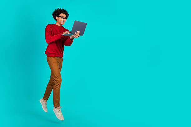 istock Young man with laptop jumping in teal studio background showcasing modern tech lifestyle and playful learning 2244132881