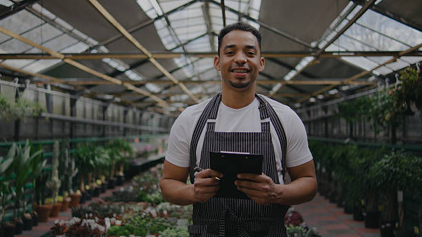 istock Man in apron stands inside a greenhouse, smiling while holding a tablet, surrounded by various plants and greenery during daytime 2244139477