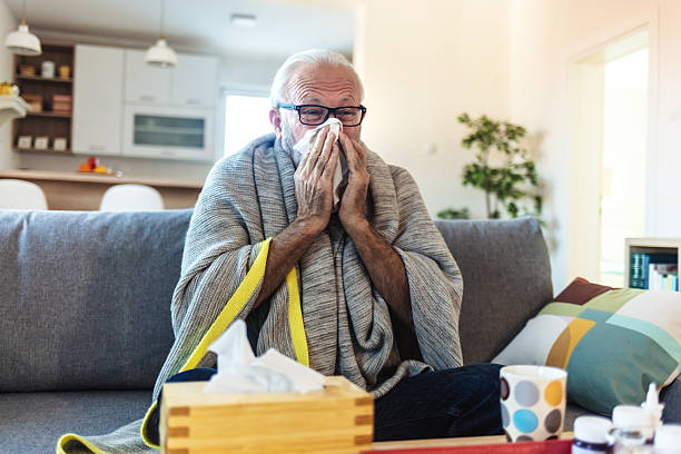 istock Man with a cold lying in sofa holding tissues. Senior man suffering from flu while sitting wrapped in a blanket on the sofa at home. Elderly Man blowing his nose while lying sick in bed at home. 2244142262