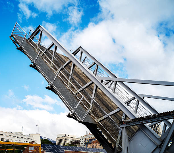Tasmania, Australia. November 23, 2019. Low-angle view of a raised steel drawbridge with truss supports and railings, set against a bright blue sky with scattered clouds.