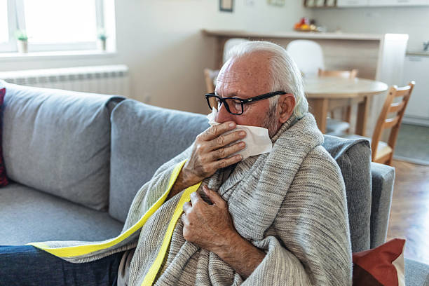 istock Man with a cold lying in sofa holding tissues. Senior man suffering from flu while sitting wrapped in a blanket on the sofa at home. Elderly Man blowing his nose while lying sick in bed at home. 2244142707
