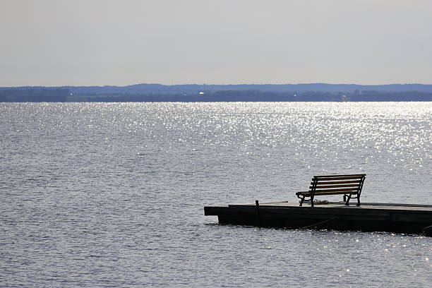 A single wooden bench sits at the edge of a small pier, facing a wide lake where sunlight sparkles across gentle ripples. The distant shoreline forms a soft, hazy line beneath a pale sky, creating a calm and contemplative scene.