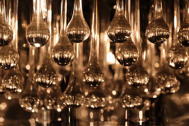Close-up of a chandelier made of elongated glass teardrops glowing with warm golden light. Reflections shimmer across the smooth surfaces, while a shallow depth of field creates soft bokeh and a rhythmic, symmetrical pattern of sparkling drops.