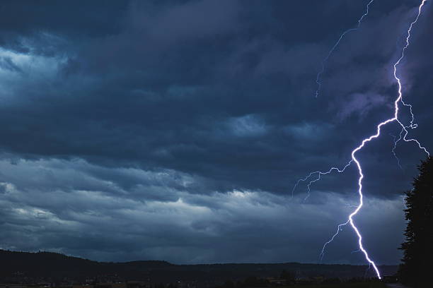 istock Lightning bolt discharging  towards urban are grounds near tall tree during thunderstorm at night. Wide angle view, dramatic cloudy skies, no people 2244155881