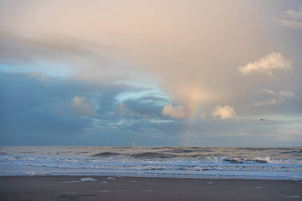 A faint rainbow glows through pastel clouds over the rolling sea. Gentle waves meet the quiet shore in soft evening light.