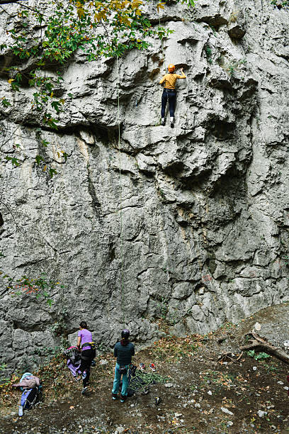Climbers tackle a rugged rock wall with safety gear as friends and mentors provide support from ground level, capturing teamwork, adventure, and outdoor sport in a forest environment.