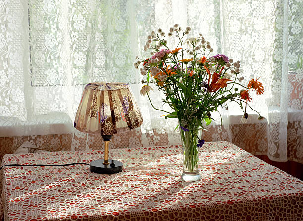 Glass vase holding assorted wildflowers standing on table next to small decorative lamp, sunlight filtering through lace curtains in background creating intricate shadow patterns