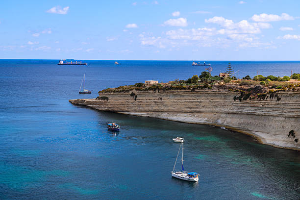 A serene coastal view of sailboats anchored near a limestone cliff under a clear blue sky. The turquoise waters gently blend with the deep blue of the open sea, creating a peaceful Mediterranean scene that captures the beauty of nature and maritime leisure.