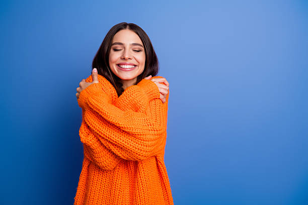 istock Joyful young woman embracing herself in a vibrant orange sweater against a solid blue backdrop expressing warmth and happiness 2244168529