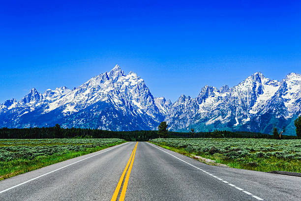 An empty highway stretches perfectly straight toward the snow-capped Teton Range under a flawless cobalt sky. The bold yellow center line divides the vast scene, drawing the eye toward the monumental peaks that rise like guardians of the American West. Fields of sagebrush flank the road, glowing in summer light, while the crisp alpine air gives each ridge a sculpted presence. This photograph symbolizes both motion and stillness — the infinite promise of the open road meeting the immovable grandeur of the mountains ahead.