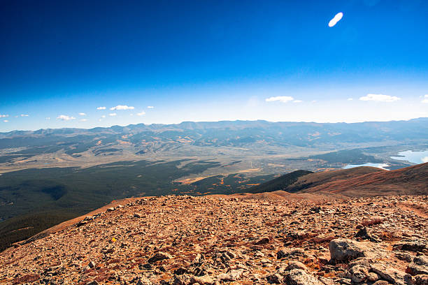 A wide, elevated view from a rocky mountain ridge captures the vast alpine valley below, dotted with forested slopes, winding terrain, and shimmering high-altitude lakes. The warm, sunlit foreground transitions into deep greens and blues across the expansive distance, while the horizon stretches under a perfectly clear sky. The composition evokes solitude, scale, and perspective — a visual embodiment of Colorado’s high country wilderness and the serenity of untouched elevation.