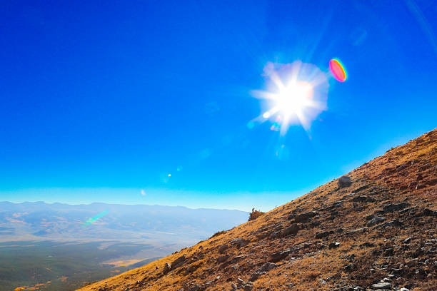 A radiant mountain landscape captured under a vivid blue sky, with the sun bursting at the frame’s edge and creating a striking lens flare across the scene. The steep, rocky slope glows with warm autumn tones, overlooking a vast valley and distant mountain ranges fading into a soft atmospheric haze. This image embodies the high-altitude brilliance and crisp clarity of the American West — ideal for editorial, fine art, or nature photography collections celebrating light, landscape, and scale.