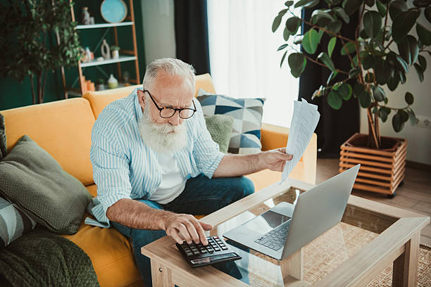istock Retired senior man with white beard works from home on a laptop while reviewing documents on a cozy living room couch 2244171016